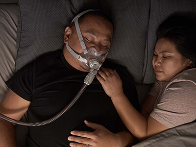 A man sleeping while wearing a ResMed CPAP mask, with his wife beside him in bed