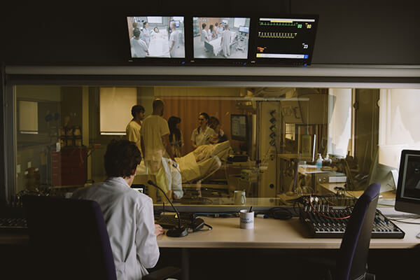 A healthcare professional in a sleep clinic control room observing a sleep apnoea diagnostic test through a glass panel.
