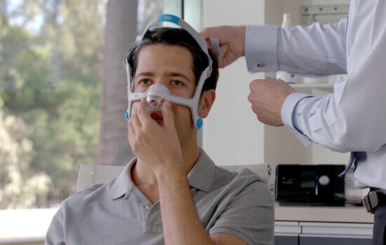 A man holding his APAP mask in place at the nose while it is fitted in a sleep clinic.
