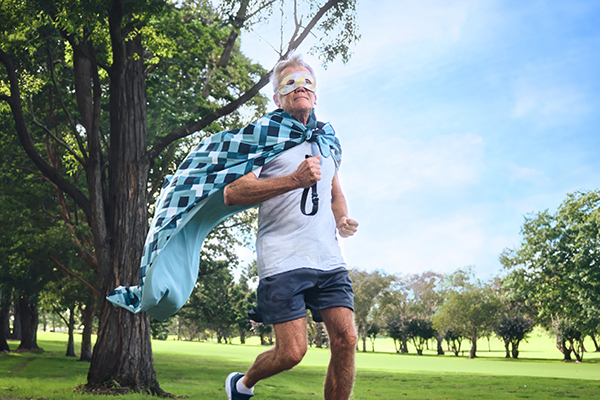 An older man running in the park wearing a superhero mask and cape made from a bed sheet, feeling energised thanks to the benefits of good sleep.