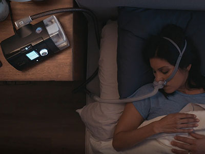 A aerial view of a woman sleep in bed wearing a CPAP mask connected to a CPAP device beside her.