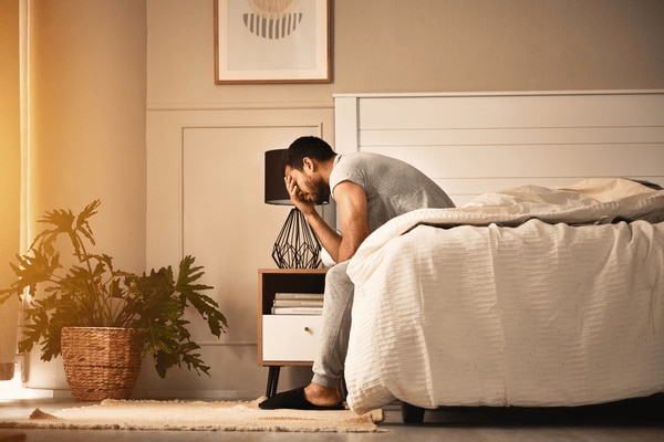  man sitting on edge of bed, exhausted from a poor night of sleep